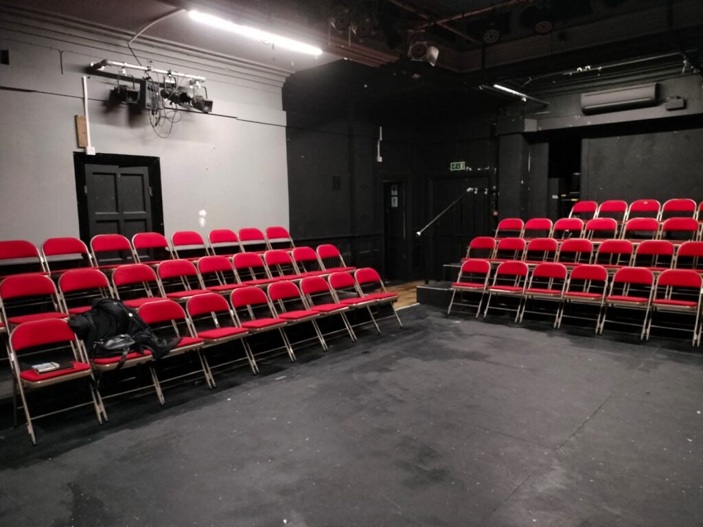 Black box theatre with red folding chairs in thrust configuration and overhead lighting rig, available for hire by the hour on Peerspace