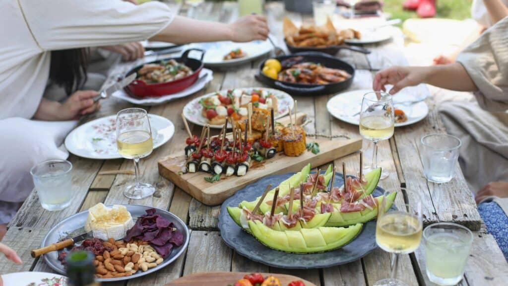 A group of people sit around a table in an outdoor setting and share plates during a summer cookout.