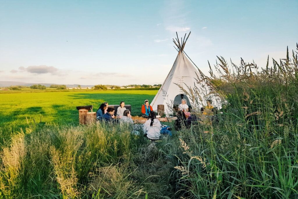 Group of women sitting in front of a tipi in a circle. Wide grass field on  a sunny day.