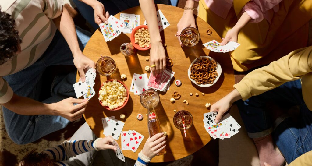 A group is sitting around a table playing a card game while eating snacks and drinking iced beverages.
