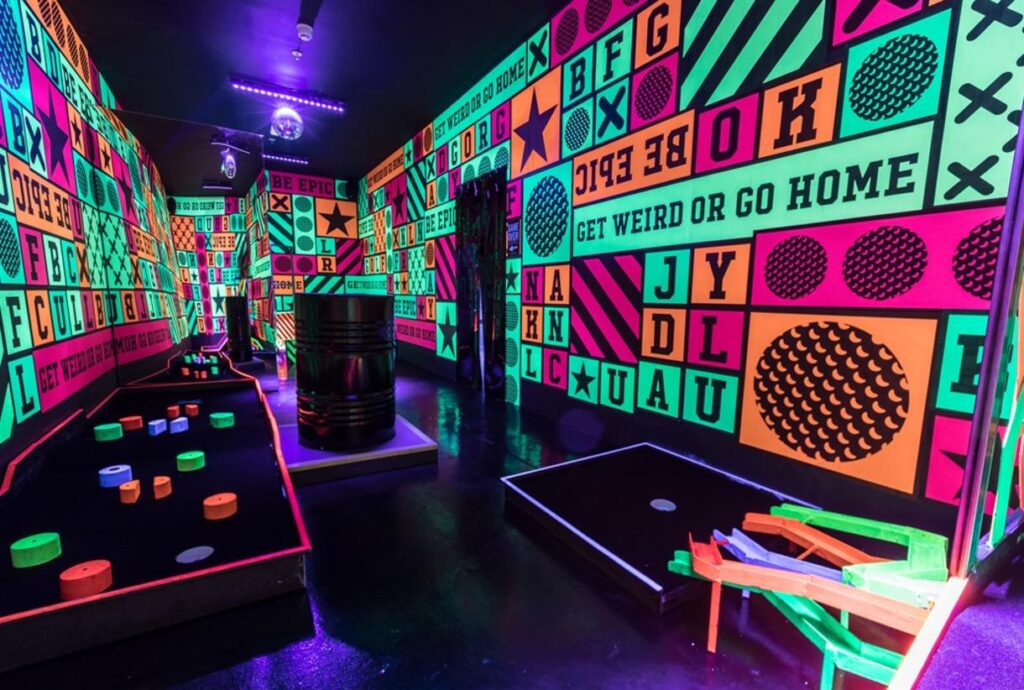 UV-lit neon mini golf course at Junkyard Golf Club with "Get Weird or Go Home" wall graphics
