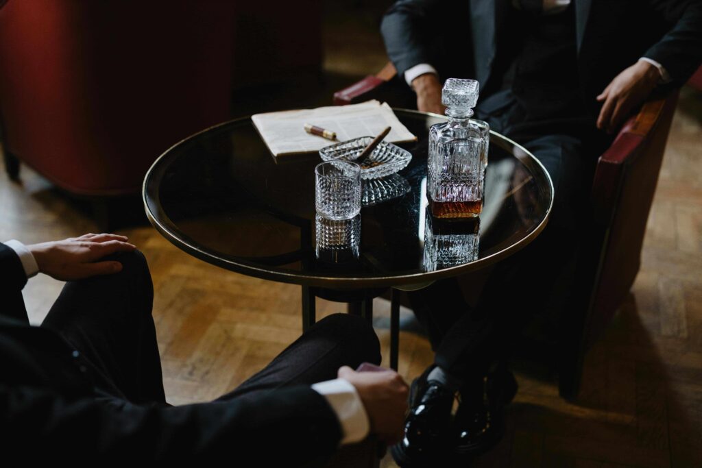 Two men in suits sitting on a bar table, drinking whisky and smoking