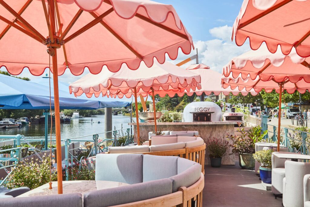 View of a sun-lit patio with half-moon seating around round tables, pink umbrellas and river views.