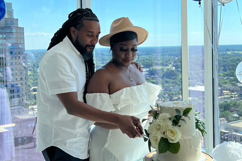 A couple standing in a brightly lit penthouse with views of the surrounding area, cutting a tiered, white cake.