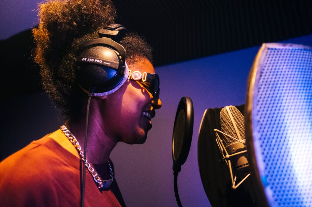 Woman singing into a microphone in a recording studio booth.