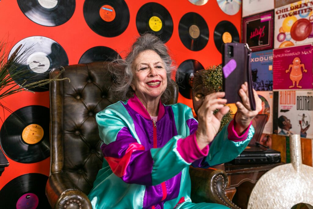 A woman in retro clothing is taking a smartphone selfie while sitting on a vintage armchair that's positioned in front of a wall decorated with vinyl records.