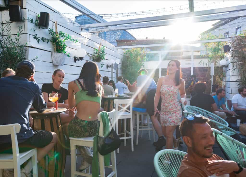 People socializing at a rooftop rental bar with white walls, plants, and sunlight streaming through a glass roof.