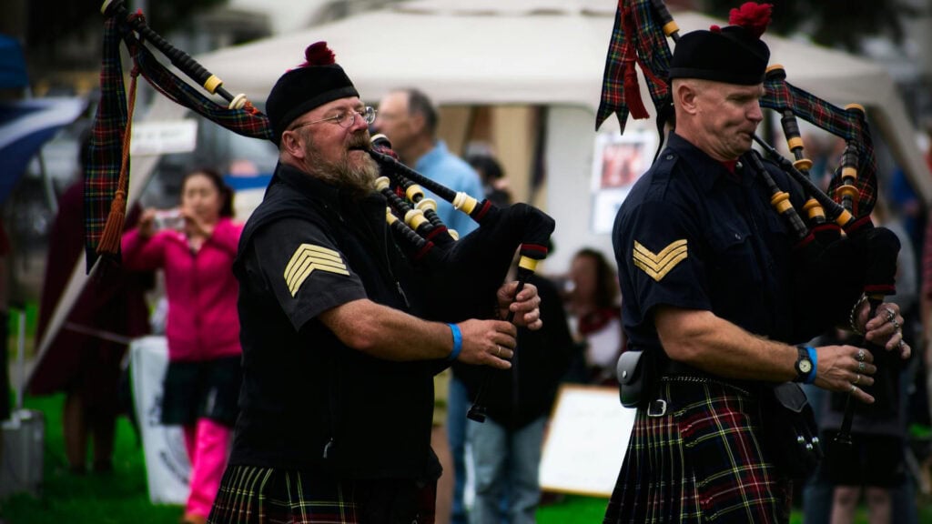Two musicians in traditional Scottish attire playing bagpipes at an outdoor event.