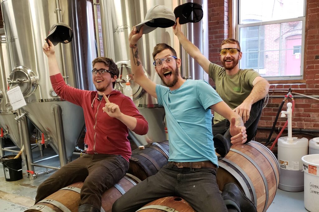 Three men sitting on beer barrels inside a brewery
