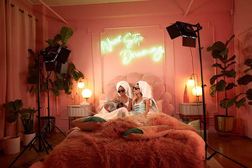 Two girls with hair towels and sunglasses sit on a bed for a photoshoot.