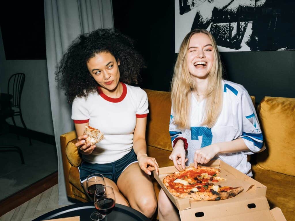 Two women eating pizza on a sofa while drinking wine.
