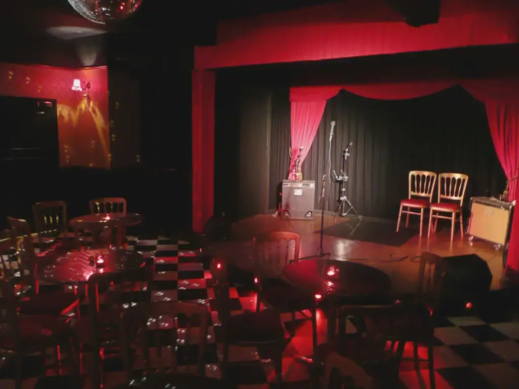 Intimate cabaret venue with red curtained stage, microphone stands, round tables and chequered floor, available for hire by the hour on Peerspace