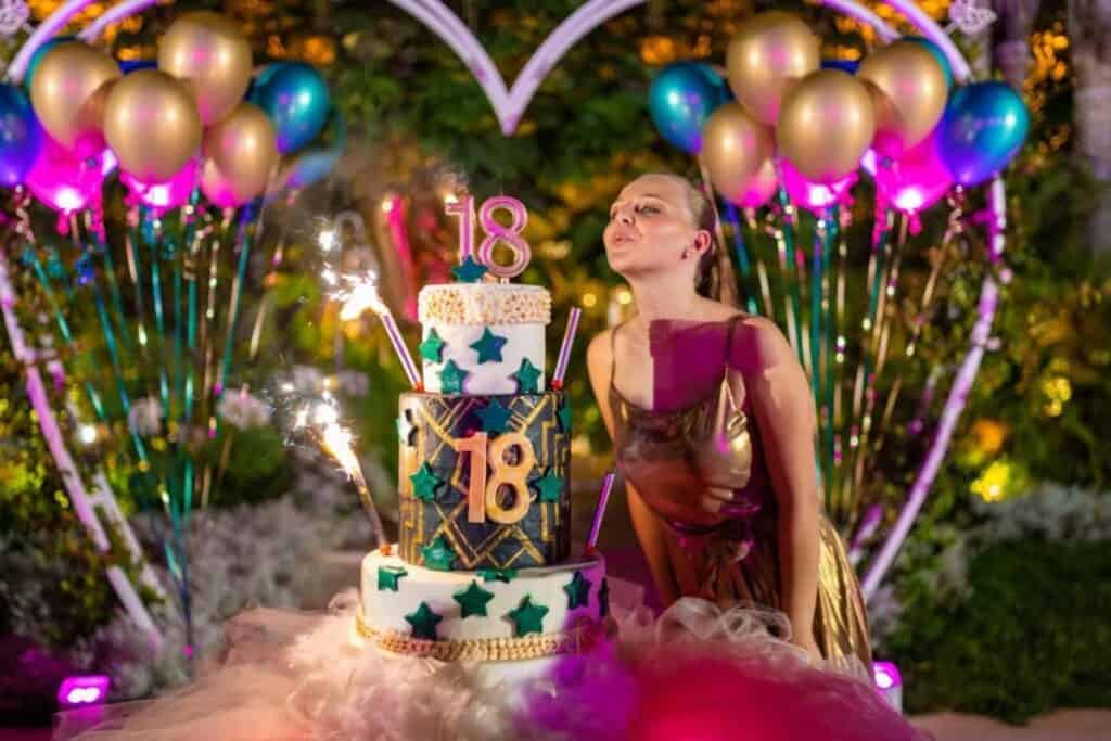 person blowing out 18th birthday cake candle with colourful balloons in the backgound