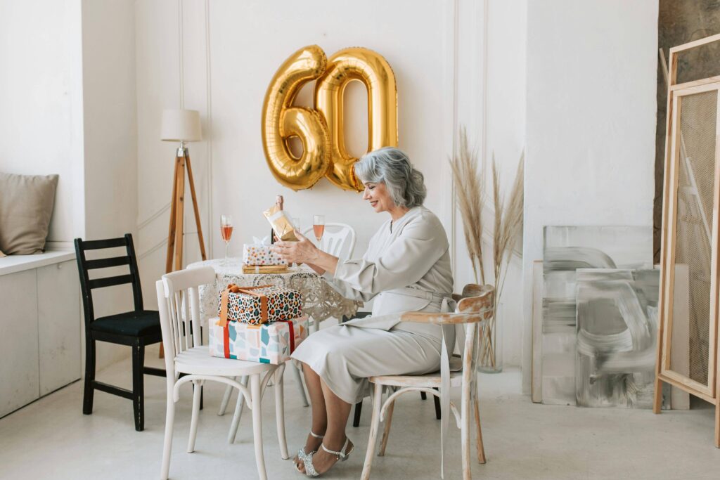 A woman sitting on a table opening her birthday gifts, with "60" balloons in the backdrop.